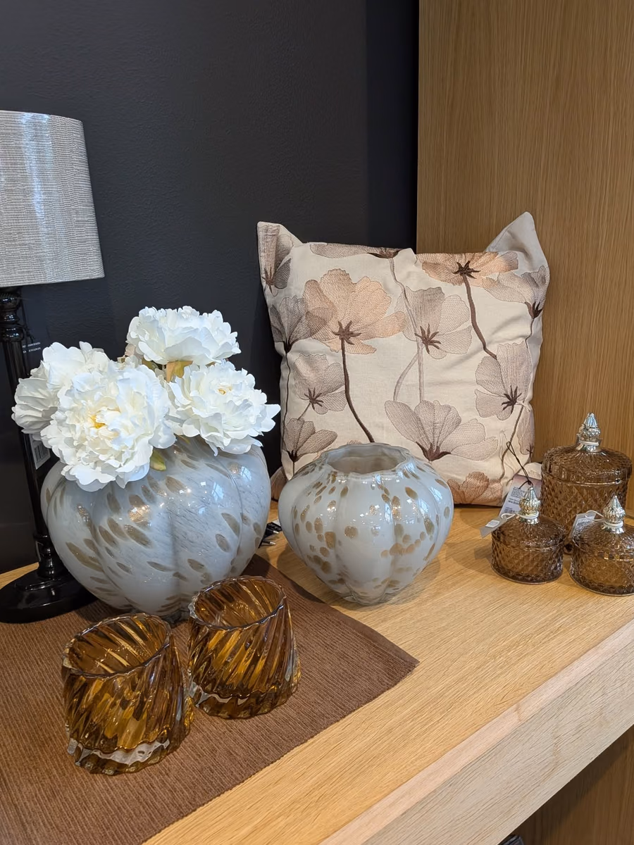 Pute med blomstermønster, brune glassvaser og lyse kunstblomster på en hylle