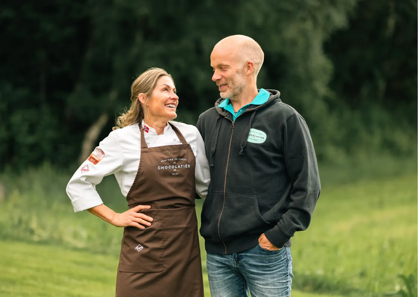 Iris i kokkejakke og forkle og Eivind i mørk genser, utendørs foran en grønn eng