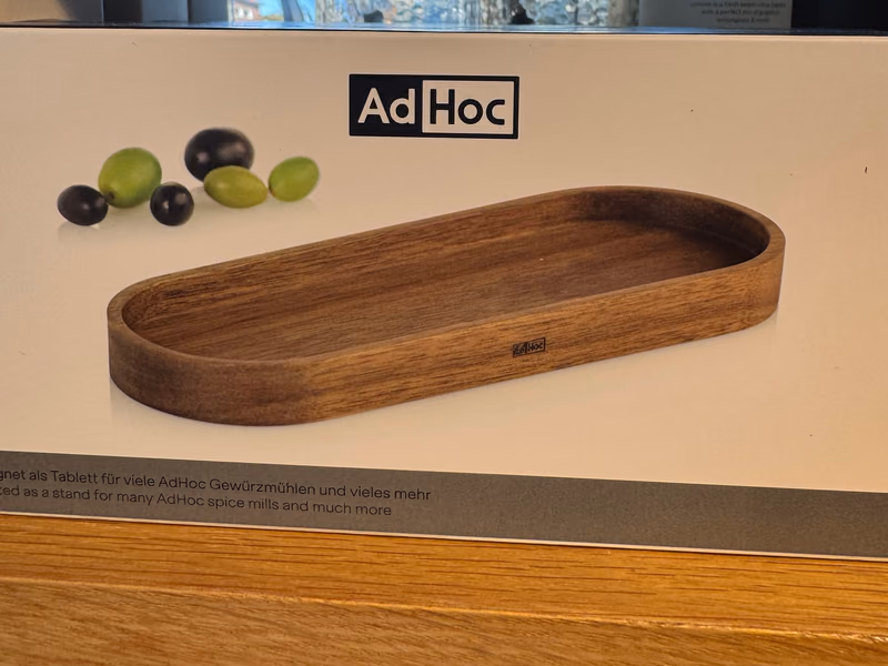 Eske med AdHoc-brett i akasie, avlangt med runde ender, avbildet på esken sammen med noen oliven, hos Gressvik Gullsmed
