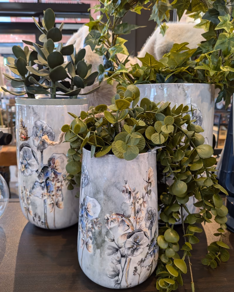 To høye krukker med malte blomster i grått og blått, med grønne planter, på et bord i interiøravdelingen hos Gressvik Gullsmed