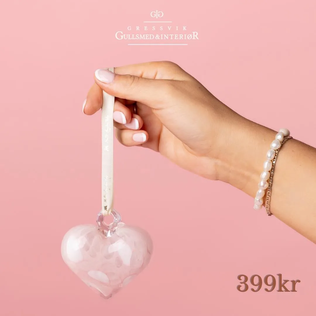 En hånd holder et rosa glasshjerte etter silkebåndet, med et perlearmbånd rundt håndleddet