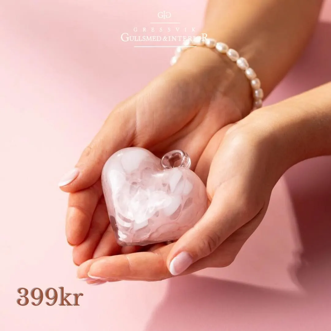 Et rosa glasshjerte med hvite skyer i glasset ligger i to hender, på rosa bakgrunn